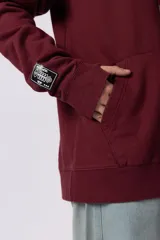 Buzo canguro color bordó con capucha y cordones ajustables. Presenta un bolsillo frontal tipo canguro y un pequeño parche rectangular con diseño gráfico en la manga derecha.