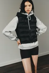 Chaleco acolchado de color negro con cuello subido, capucha ajustable y cierre frontal con cremallera termosellada. Presenta un diseño con paneles laterales en tejido liso y bolsillos laterales.