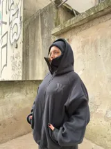 Campera oversize negra de felpa suave con cierre frontal, doble capucha que incluye un diseño tipo balaclava integrado, bolsillos laterales y logo bordado tono sobre tono en el pecho.