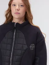 Campera crop de corte boxy y color negro, forrada en corderito, con parches capitoneados en el pecho y las mangas. Cuenta con cierre frontal metálico, cuello alto, puños y bajo elastizados, y detalle de logo bordado en hilo en el pecho.