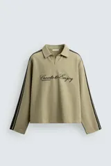 Sudadera de corte boxy fit color verde oliva, con cuello solapa y abertura frontal. Presenta mangas largas con bandas laterales negras a contraste y bordado frontal con texto en color negro.