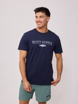 Remera azul marino de manga corta con estampado frontal que incluye el texto "Rusty Supply Surfboards" y el logo de la marca.