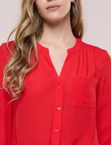 Blusa de calce relajado color salmón, confeccionada en tela liviana con textura sutil. Presenta escote en V con abotonadura frontal, bolsillo parche en el pecho, mangas largas regulables con presilla y ruedo con opción de anudar en el frente.