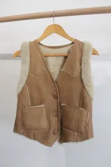 Chaleco de cuero genuino color tostado con interior y detalles en piel sintética color crema. Presenta cierre frontal con botones, dos bolsillos delanteros y diseño con costuras visibles.