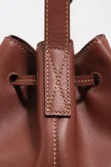 Bolso tipo saco de cuero marrón con cierre de cordón ajustable, asa larga regulable y logotipo de BIMBA Y LOLA impreso en el frente con letras blancas.