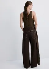 Pantalón de jean color marrón oscuro, de corte recto y tiro medio. Presenta una hilera de tachas metálicas plateadas a lo largo de la costura lateral externa.