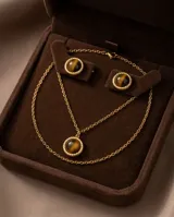 Set compuesto por un collar con cadena fina y dije circular, y un par de aros tipo botón a juego. Ambas piezas presentan una piedra natural ojo de tigre engarzada en una estructura de metal dorado con borde trenzado.
