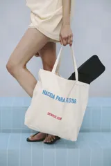 Bolso tipo tote de tela color crema con asas largas. Estampado frontal con la frase "NACIDA PARA FLOTAR" en azul y "SRTAPEEL.COM" en rojo.