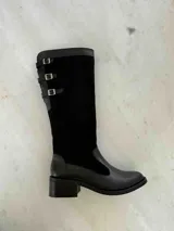 Bota alta de cuero color marrón chocolate, con taco cuadrado de 5 cm y caña alta hasta la rodilla. Presenta tres correas con hebillas en la parte superior de la caña. Fabricada en Colombia.