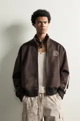 Cazadora relaxed fit de tejido efecto ante color marrón oscuro, con cuello subido y cierre frontal de cremallera. Presenta mangas largas con bandas blancas a contraste en los hombros, bolsillos laterales de vivo, y bordados en hilo blanco en el pecho.