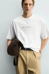 Camiseta blanca de corte relajado, con cuello redondo, manga corta y estampa frontal de texto pequeño en marrón.