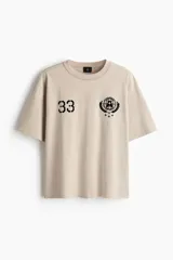 Remera color beige de punto pesado de algodón, de corte holgado, cuello redondo ribeteado y hombros caídos. Presenta estampados en negro en el pecho: el número "33" a la izquierda y un emblema circular con estrellas a la derecha.