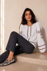 Blusa blanca de manga larga con detalles de bordados florales en tono grisáceo y bandas decorativas en el frente y mangas. Cuenta con cuello redondo y puños elásticos.