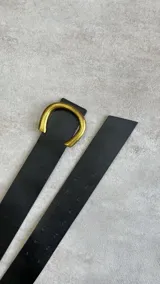 Cinto negro de cuero sintético con hebilla dorada en forma de letra C.