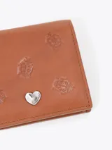 Mini billetera de cuero vacuno color rojo, con diseño grabado de pequeñas mariquitas en el frente y un aplique metálico en forma de corazón. Cuenta con un interior funcional con espacios organizadores para tarjetas.