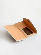 Billetera de cuero vacuno color crudo con textura granulada. Presenta un diseño tipo sobre con cierre de broche y un detalle decorativo de espiga bordado en cuero color suela en la solapa frontal.