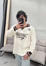 Remera tejida de manga larga con cuello polo marrón, diseño perforado y estampado frontal con la frase "ENDURING Courage".