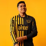 Camiseta de fútbol Puma del Club Atlético Peñarol, edición aniversario, modelo de manga larga. Diseño con mitad derecha a rayas verticales amarillas y negras, y mitad izquierda negra con escudo del CURCC y logo de Puma en dorado. Presenta cuello polo negro y sponsor antel en el pecho.