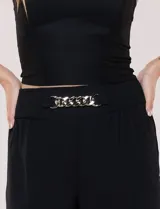 Pantalón negro de calce relajado y caída fluida, confeccionado en poliéster con textura de crepé. Tiene cintura alta elastizada estilo pull on con un aplique de cadena metálica decorativa, y pierna de corte ancho tipo wide leg corto por arriba del tobillo.