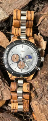 Reloj cronógrafo con caja de madera de zebrano y bisel técnico de acero negro. La esfera es plateada con subdiales de madera, indicador de fase lunar y fechador. La malla combina eslabones de acero inoxidable con madera de zebrano.