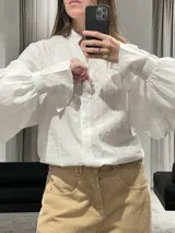 Camisa blanca de textura rayada con cuello mao y cierre frontal de botones. Presenta mangas largas abullonadas con puños anchos y corte holgado.