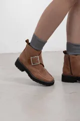 Botines de cuero vacuno color suela con textura envejecida, suela track negra con pespunte en contraste, solapa cruzada con gran hebilla metálica plateada y cierre lateral interior.