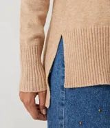 Suéter de tejido de punto color beige con cuello alto, mangas con corte raglán y puños y ruedo acanalados. Presenta un diseño clásico y cómodo con el ruedo trasero ligeramente más largo.