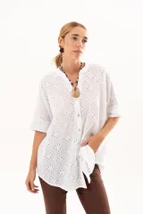 Camisa de broderie blanca con silueta oversize, escote en V, cierre frontal con botones y mangas 3/4. Presenta un diseño con bajo asimétrico, siendo ligeramente más larga en la parte posterior.