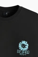 Remera de algodón negra, de corte holgado y diseño de hombros caídos. Presenta un pequeño estampado en el pecho con una flor celeste y el texto "KEEP THE WILD ALIVE".