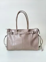 Bolso grande de cuero marrón con textura tipo cocodrilo, estructura rígida y asas para llevar al hombro. Tiene una tira con tachas y hebillas en cada extremo, también lleva lazos en los costados.