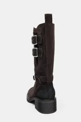 Botín de mujer de serraje color marrón oscuro, estilo motero, con caña media y tres tiras ajustables con hebillas metálicas plateadas. Posee tacón de bloque medio y tirador trasero para facilitar el calce.