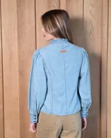 Camisa de jean blanca con cuello alto con volados, cierre frontal con botones oscuros y mangas largas abullonadas con puños ajustados.
