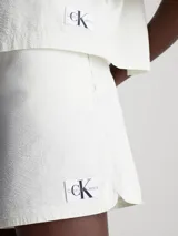 Shorts de mujer color blanco roto, confeccionados en sirsaca de algodón. Tienen un corte regular, cintura con cordón ajustable, bolsillos laterales y bolsillos traseros de ojal. Presentan una insignia con el logo de Calvin Klein en el bajo.