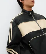 Campera estilo racing confeccionada en poliuretano, con diseño bicolor en negro y beige. Presenta cuello alto con botón a presión, cierre frontal metálico, panel frontal contrastante y franjas laterales deportivas.