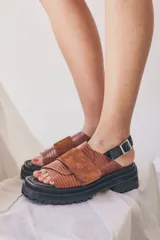 Sandalias de plataforma con suela track negra. El diseño presenta tiras cruzadas en cuero texturizado color marrón con detalles en gamuza, y una tira trasera ajustable con hebilla metálica.