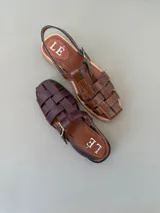 Sandalias de cuero marrón con diseño tipo huarache de tiras entrelazadas, talón abierto con tira ajustable y hebilla metálica.