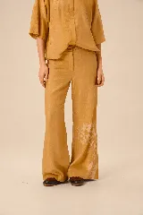 Pantalón de lino color mostaza, de corte wide leg, con sutil estampado floral en la parte inferior de la pierna izquierda y botón a la vista en la cintura.