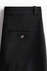 Pantalón negro de corte bootcut, con tiro alto, trabillas para cinturón y cierre frontal con botón y cremallera. Cuenta con pliegues marcados al frente y bolsillos simulados con solapa en la parte delantera.
