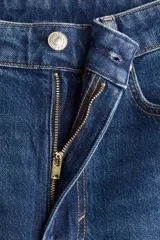 Jean de tiro alto con corte bootcut, confeccionado en denim de algodón con leve elasticidad. Presenta diseño clásico de cinco bolsillos, cierre frontal con botón y perneras ajustadas desde la cintura hasta el muslo, ensanchándose ligeramente desde la rodilla hacia el bajo.