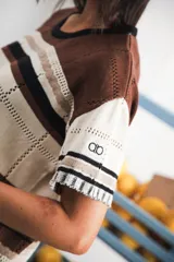 Vestido largo de tejido calado con rayas horizontales en tonos beige, marrón y amarillo. Tiene corte recto, hombros caídos, tajo lateral y logo bordado en la manga.