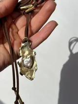 Collar artesanal con dije de base metálica irregular y acabado texturizado, que presenta una perla central incrustada. El dije está suspendido de un tiento de gamuza marrón con detalles de terminales metálicos cilíndricos.