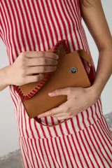 Cartera de hombro pequeña de cuero color tostado, con solapa y detalles de bordado artesanal en hilo rojo. Cuenta con correa ajustable y cierre con broches imantados.