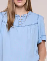 Blusa celeste de lyocell ultraliviano, de estilo romántico y silueta holgada. Presenta escote redondo con abertura en V y botones frontales, canesú fruncido y mangas cortas amplias.
