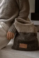 Cartera tipo tote tejida a mano en lana, con diseño bicolor en tonos beige y marrón. Presenta una etiqueta de cuero con el logo de la marca en el frente.