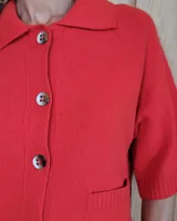 Cardigan de punto color arena, con cuello camisero, cierre frontal de botones y mangas cortas con terminación acanalada. Presenta dos bolsillos frontales tipo parche.