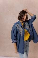 Camisa de denim azul con bordados florales en tonos amarillos y celestes sobre los hombros y el pecho. Presenta cuello clásico, cierre frontal con botones y mangas largas con puños.
