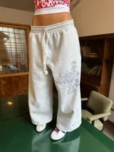 Pantalón de jogging gris de corte baggy, confeccionado en algodón. Presenta cintura elástica con cordón largo ajustable, botones en la parte inferior para ajustar la botamanga y un diseño artístico que combina estampa en plastisol con bordado en relieve sobre el muslo izquierdo.