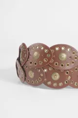 Cinturón de cuero vegano color marrón, compuesto por una serie de piezas circulares unidas, cada una con un aplique metálico central en forma de sol y detalles de tachas circulares. Incluye una hebilla metálica grande con diseño floral calado.