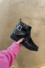 Bota corta de cuero y gamuza color negro, con taco bajo y suela plana. Presenta una correa lateral de cuero charolado con hebilla metálica, tachas decorativas en la puntera y paneles elásticos en los laterales de la caña.