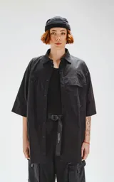 Camisa de manga corta color negro, confeccionada en popelín de algodón, con silueta oversize y dos bolsillos frontales, uno con solapa y otro con ojal y botón.